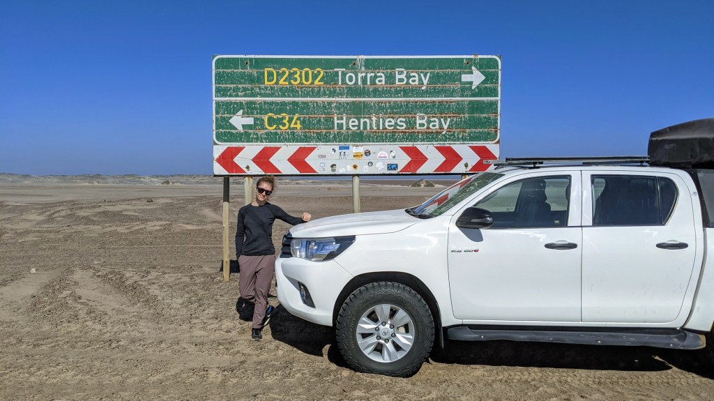 Woman standing in front of a car at at road sign on the Skeleton Coast in Namibia